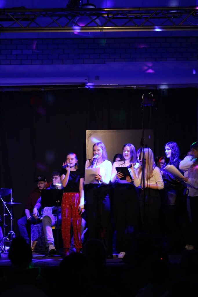 Schülerinnen singen auf einer Bühne, begleitet von einer Band. Beleuchtung ist vorhanden.