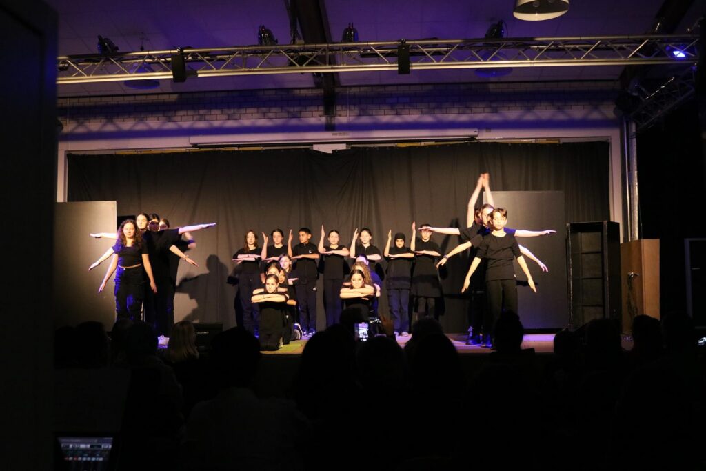 Tanzgruppe in schwarzer Kleidung führt auf einer Bühne eine Choreografie auf.