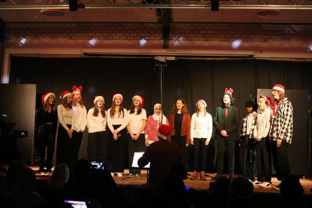 Schülergruppe singt auf einer Bühne in festlicher Kleidung mit Weihnachtsmützen.