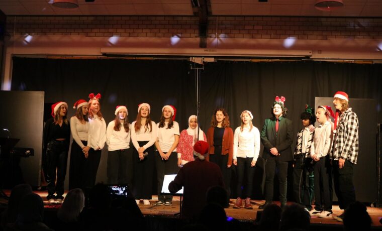 Schülergruppe singt auf einer Bühne in festlicher Kleidung mit Weihnachtsmützen.