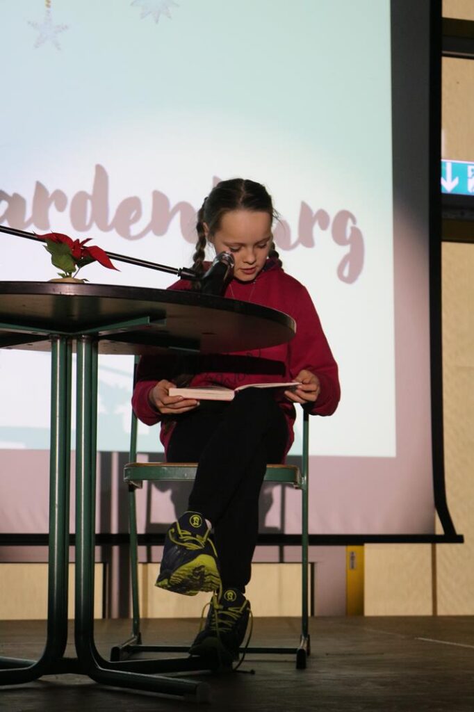 Ein Mädchen sitzt an einem Tisch und liest in ein Mikrofon.