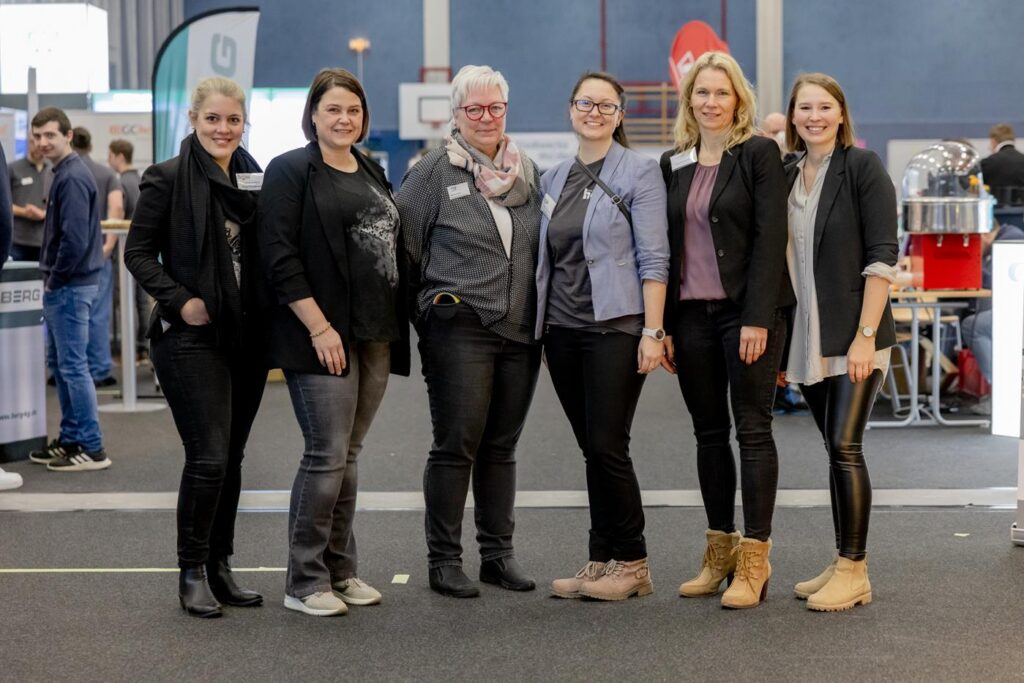 Sechs Frauen stehen in einer Ausstellungshalle, lächelnd und in unterschiedlichen Outfits.