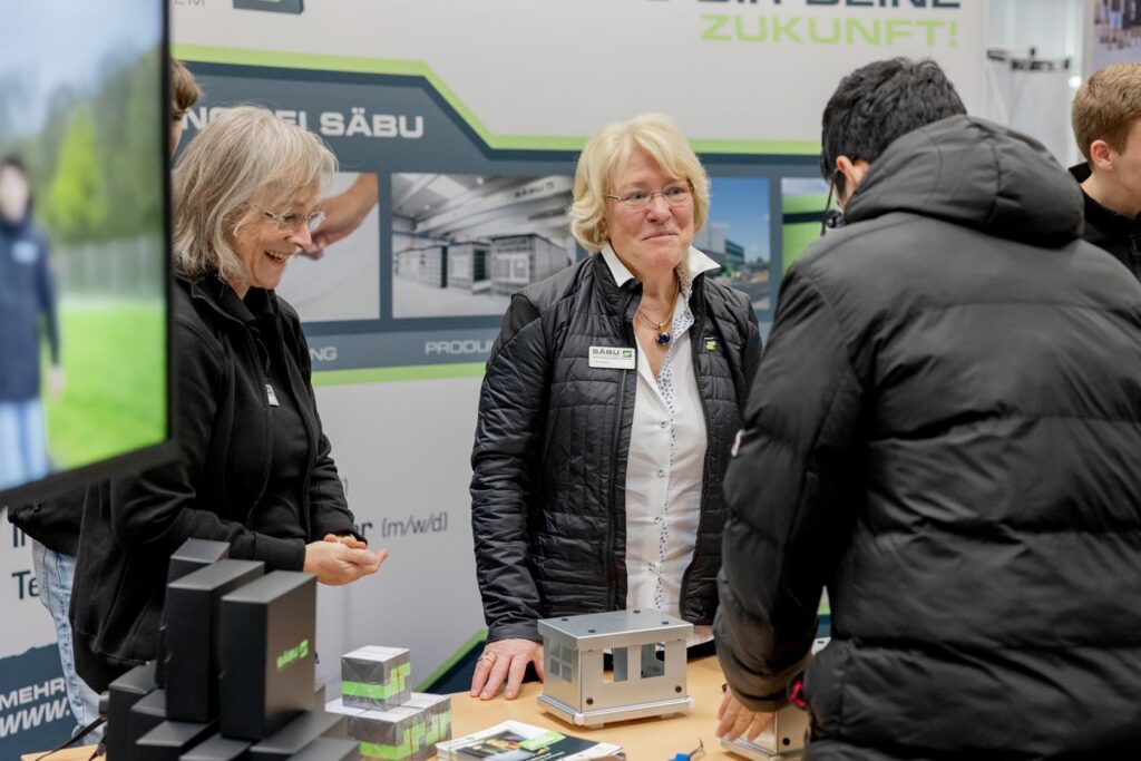 Aktiv am Messestand mit zwei Frauen und Besuchern, Materialien sichtbar.