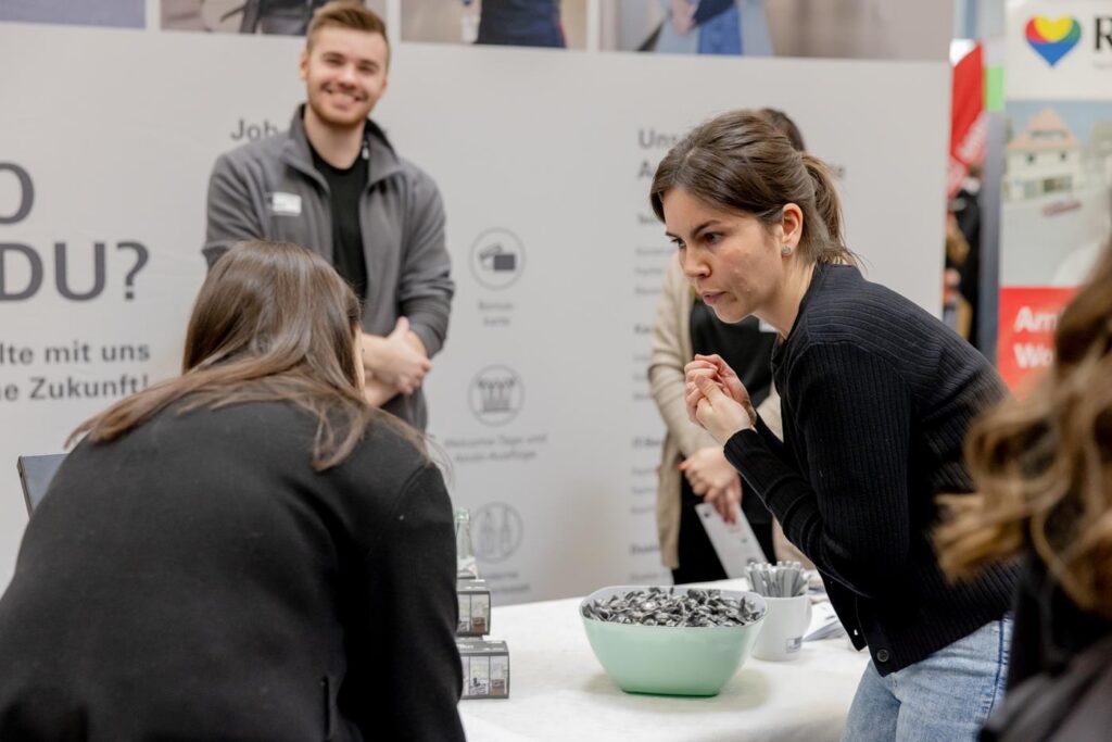 Zwei Personen am Tisch, eine zeigt Interesse, der andere lächelt am Stand.