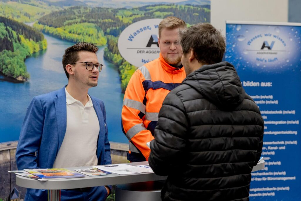 Messebesucher spricht mit zwei Standmitarbeitern an einem Informationsstand.