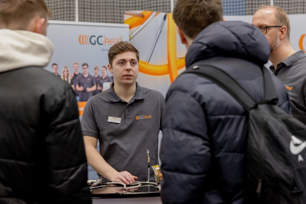 Ein junger Mann erklärt etwas an einem Messestand, Besucher hören aufmerksam zu.