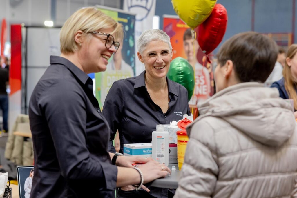 Zwei Frauen in schwarzen Shirts stehen an einem Stand und sprechen mit einer Besucherin.
