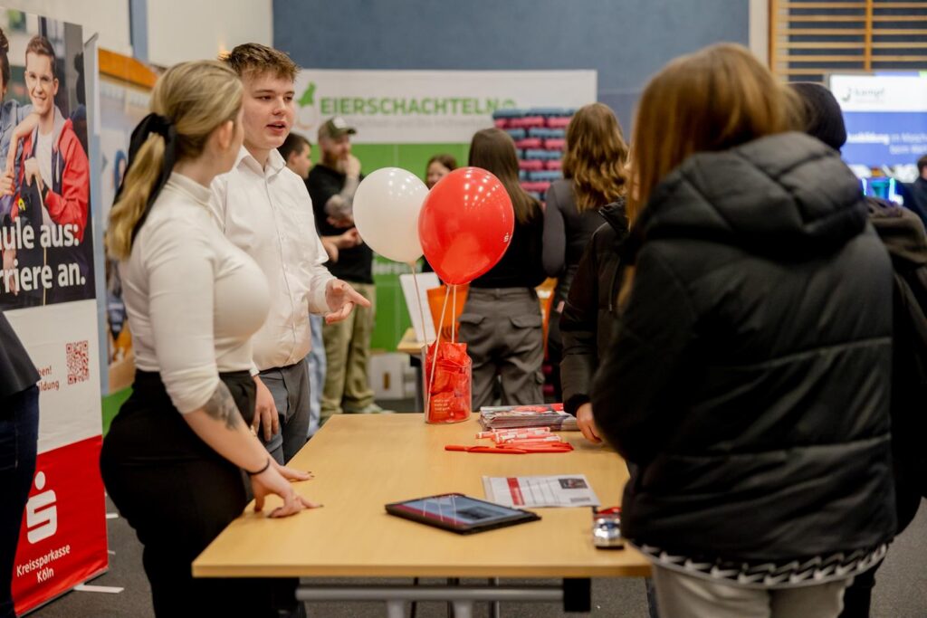 Zwei Personen sprechen am Tisch, daneben stehen Besucher und Dekoration mit Luftballons.