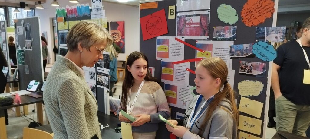 Zwei Schülerinnen präsentieren ein Projekt vor einer Besucherin auf einer Ausstellung.