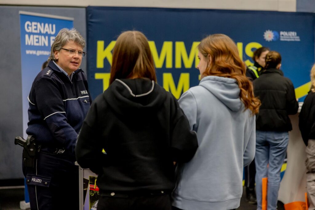 Zwei Schülerinnen stehen vor einer Polizistin bei einer Informationsveranstaltung.