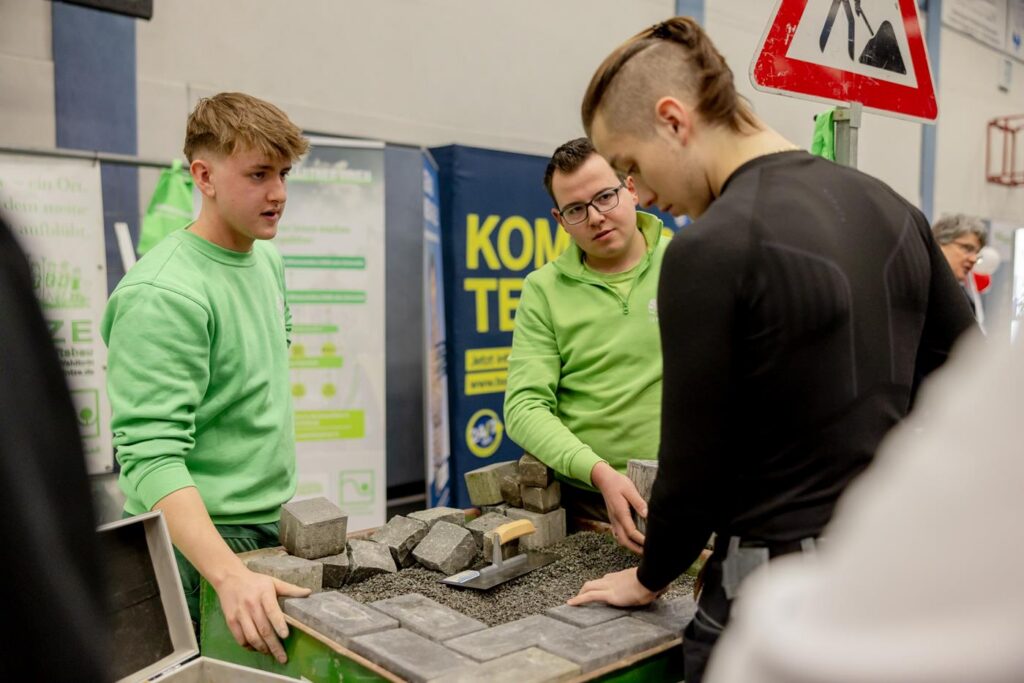 Drei Männer betrachten Baumaterialien an einem Tisch in einer Ausbildungsmesse.
