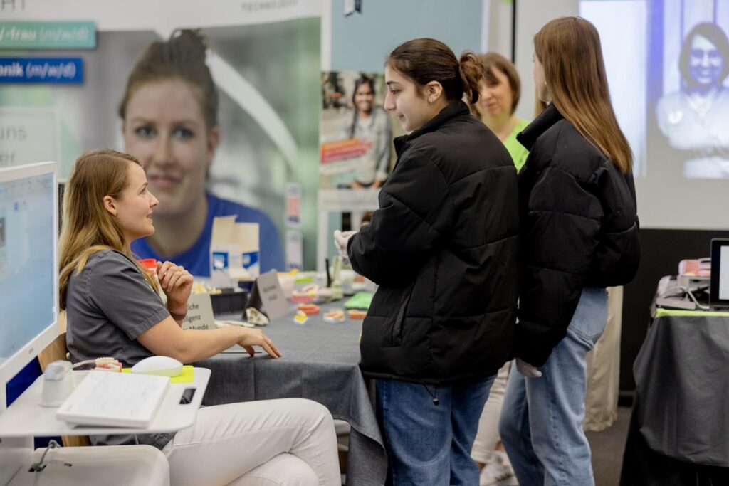 Mädchen stehen an einem Stand, während eine andere Person ihnen Informationen gibt.