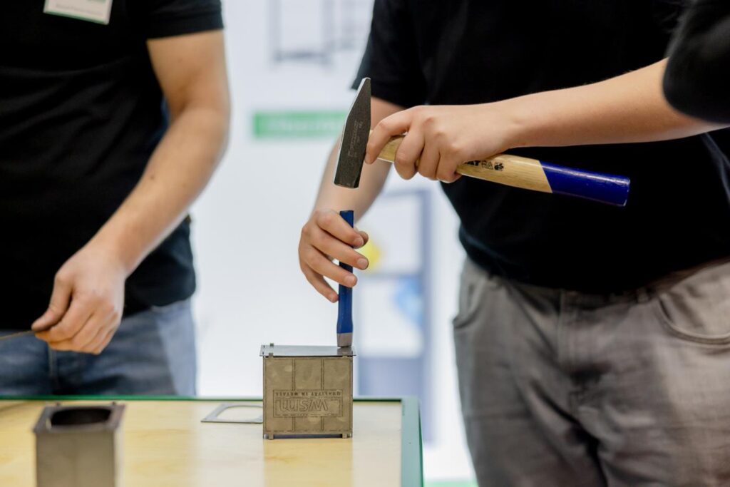 Zwei Personen arbeiten am Tisch mit Werkzeugen und einem Metallobjekt.