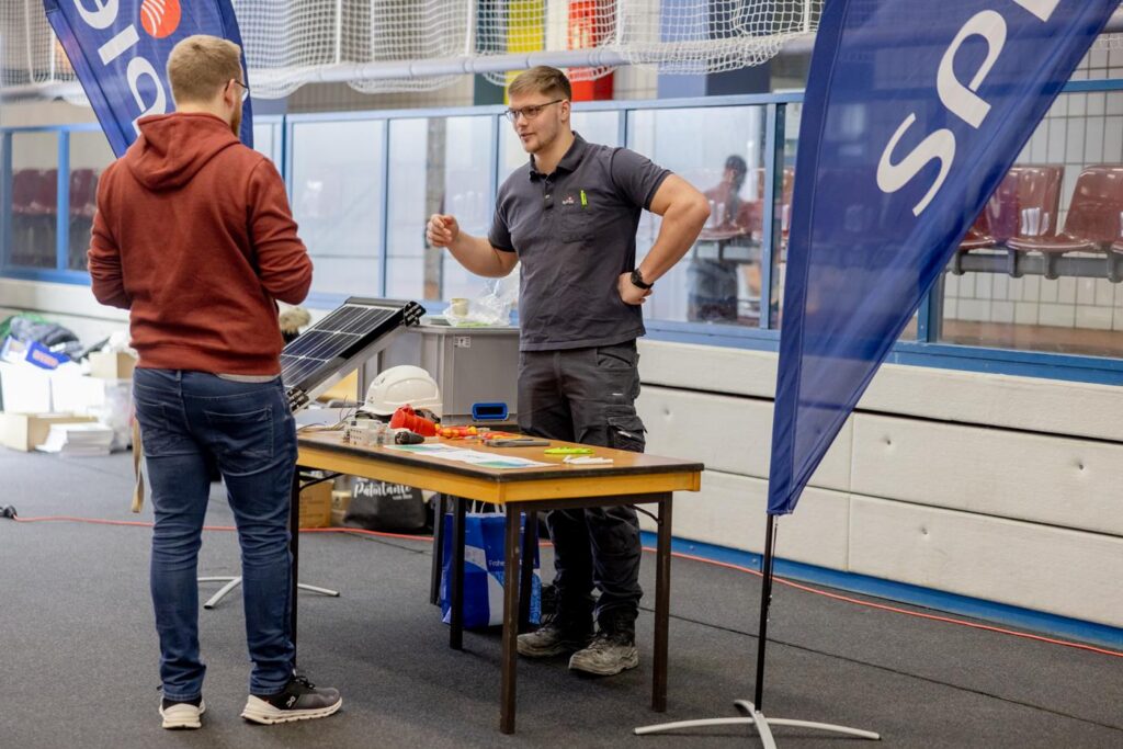Zwei Personen stehen an einem Tisch mit Solartechnologie, im Hintergrund Fahnen.