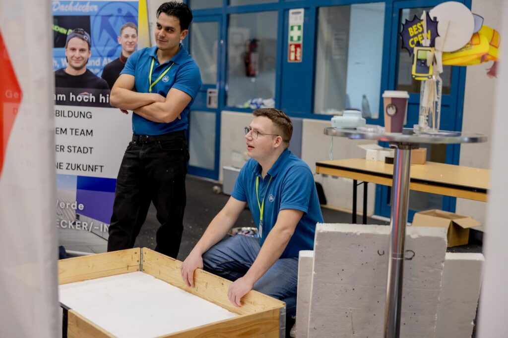 Zwei Personen in blauen Shirts an einem Stand, einer sitzt auf dem Boden, der andere steht daneben.