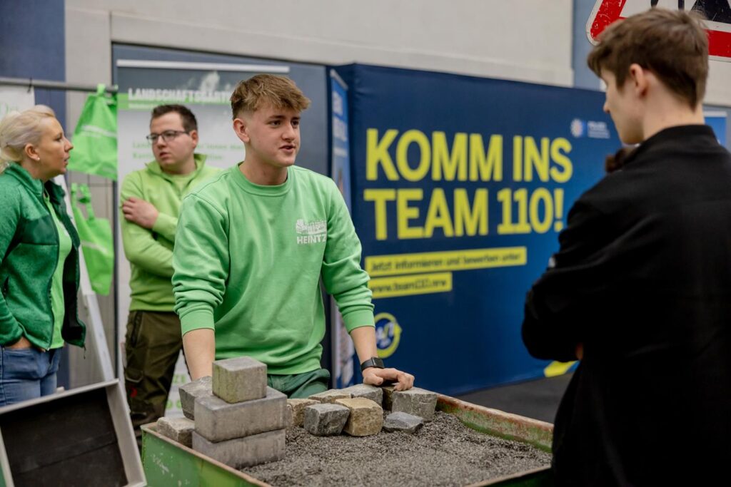 Eine Person in grüner Kleidung erklärt etwas am Stand, Steine liegen auf einem Tisch.