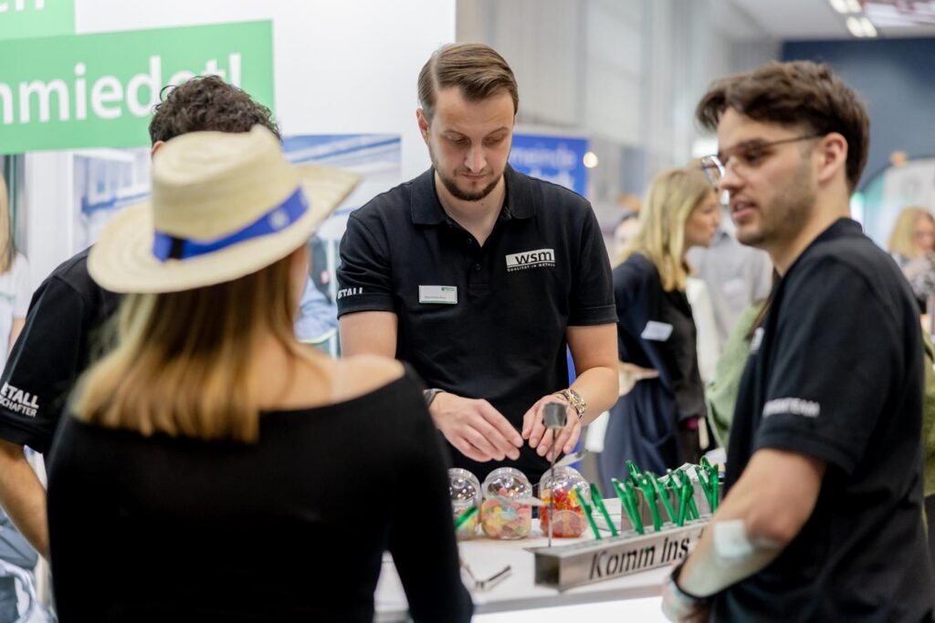 Eine Person im Hut spricht mit mehreren Personen an einem Messestand.