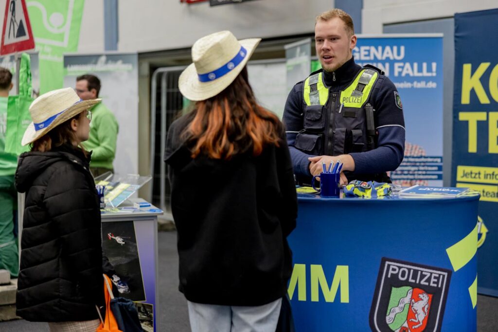 Ein Polizist steht an einem Informationsstand und spricht mit zwei Jugendlichen.