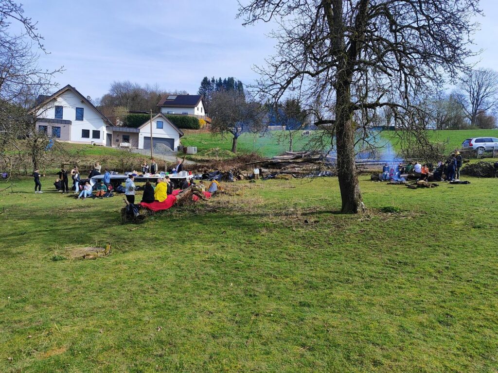 Gruppen von Menschen auf einer Wiese, einige beim Grillen, Häuser im Hintergrund.