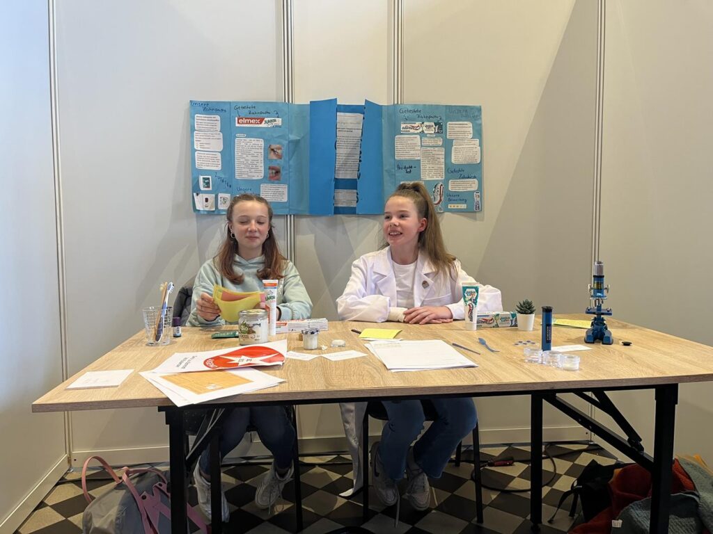 Zwei Schülerinnen sitzen an einem Tisch mit wissenschaftlichen Materialien und einem Plakat.