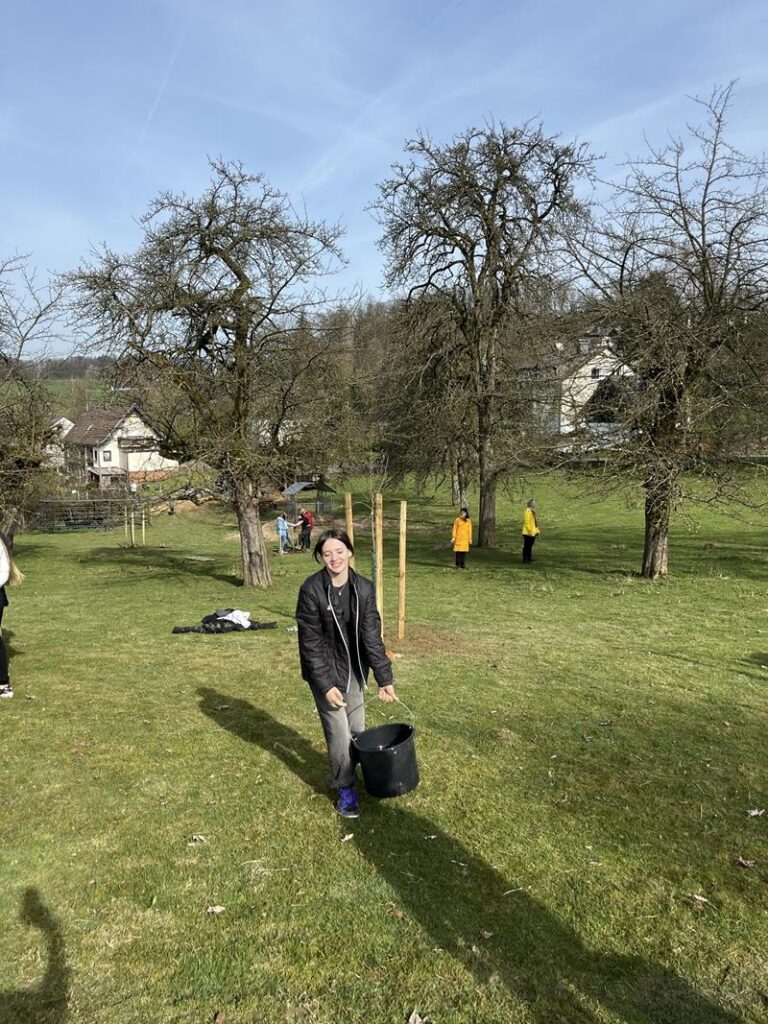 Schüler tragen Eimer auf einer Wiese mit Bäumen und Häusern im Hintergrund.