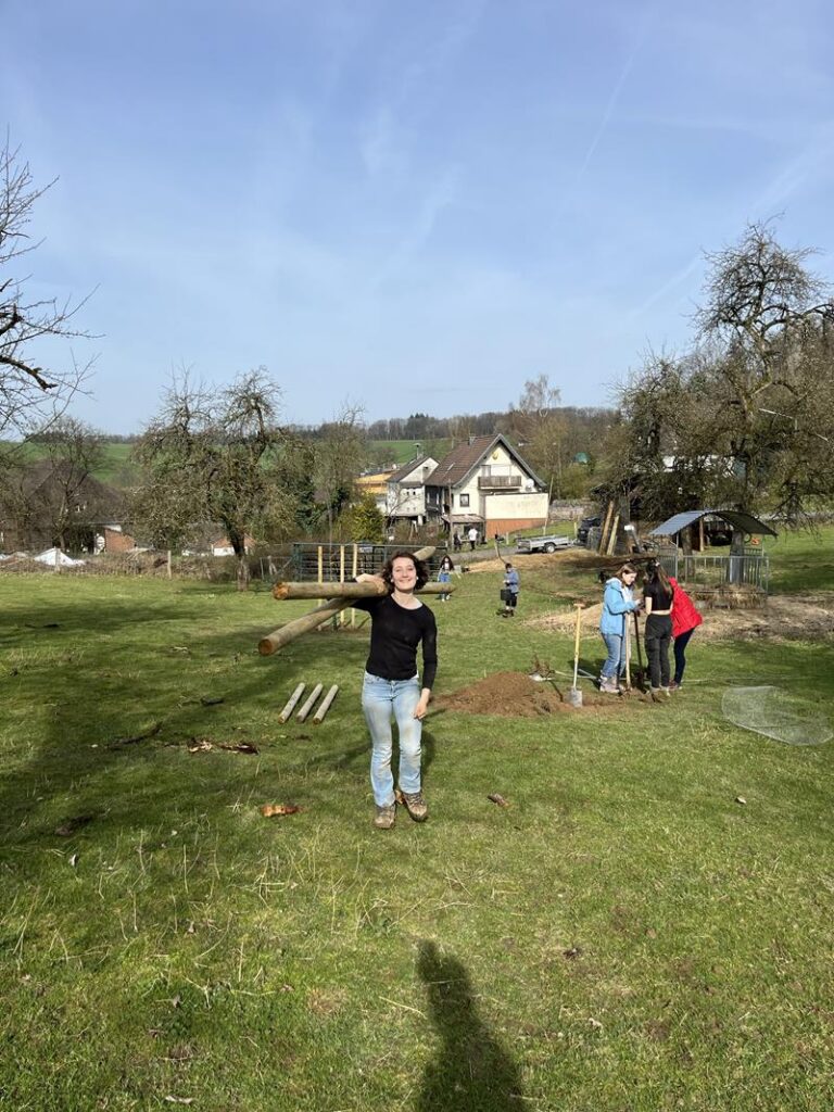 Person steht auf einer Wiese, im Hintergrund sind Häuser und weitere Personen beim Arbeiten.