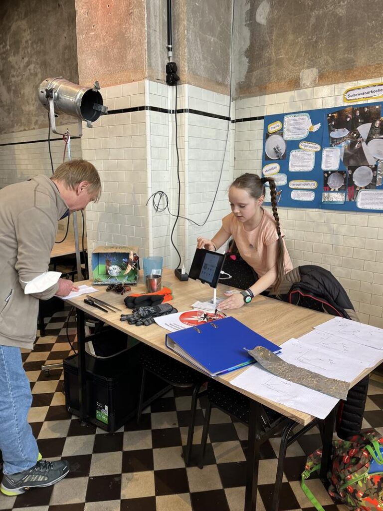 Personen arbeiten an einem Tisch in einem Raum mit Schautafeln. Ein Laptop und Materialien liegen bereit.