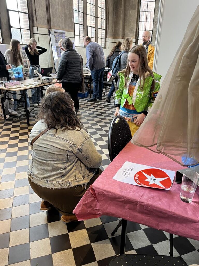 Personen in einem Raum im Gespräch, Tische mit Auslagen im Hintergrund.