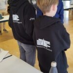 Zwei Schüler mit Kapuzenpullis stehen an einem Tisch in einem Klassenraum.