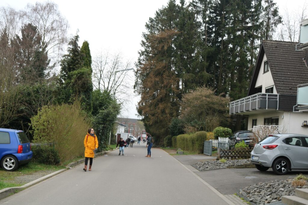 Belebte Straße mit Fußgängern und parkenden Autos, umgeben von Bäumen und Wohnhäusern.
