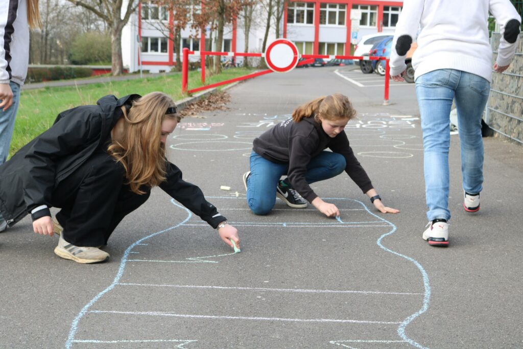 Schülerinnen zeichnen mit Kreide auf dem Gehweg.
