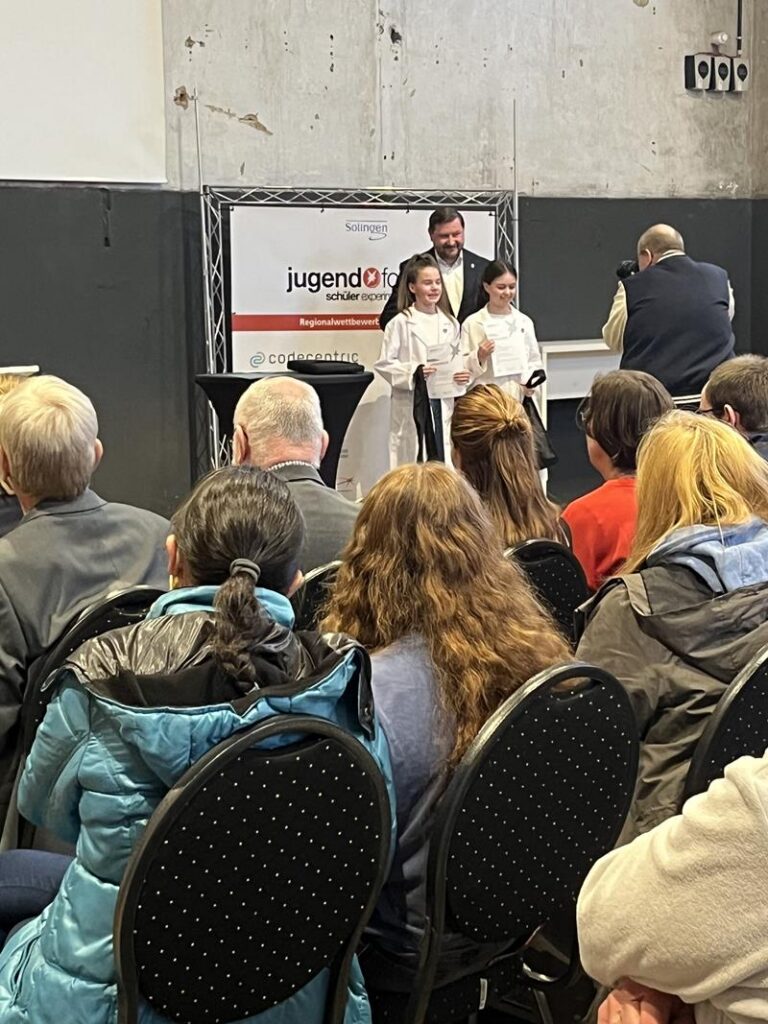 Zwei Kinder stehen auf der Bühne und präsentieren etwas vor einem Publikum.