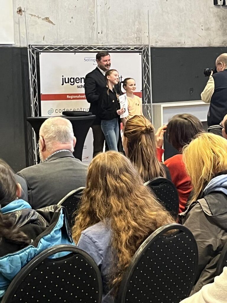 Veranstaltung mit Publikum, Vortragender und zwei Personen auf der Bühne.