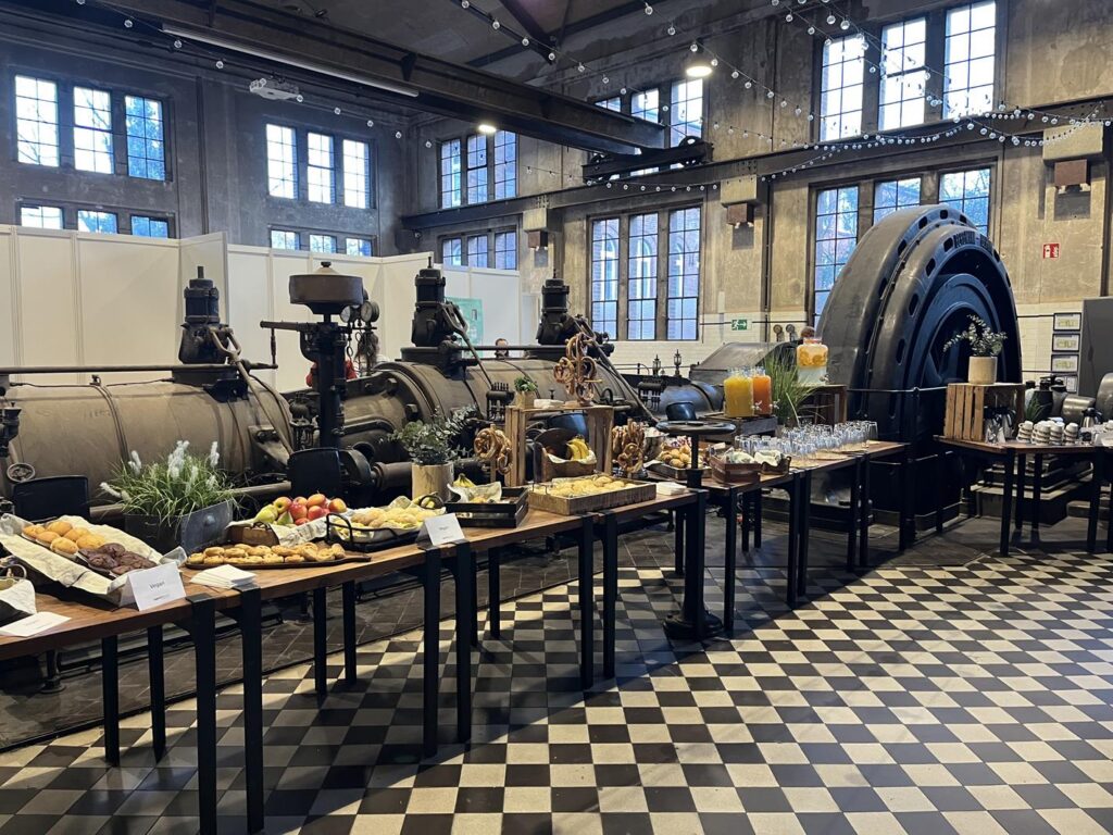 Buffet mit Speisen und Getränken in einer Industriehalle mit großen Maschinen im Hintergrund.