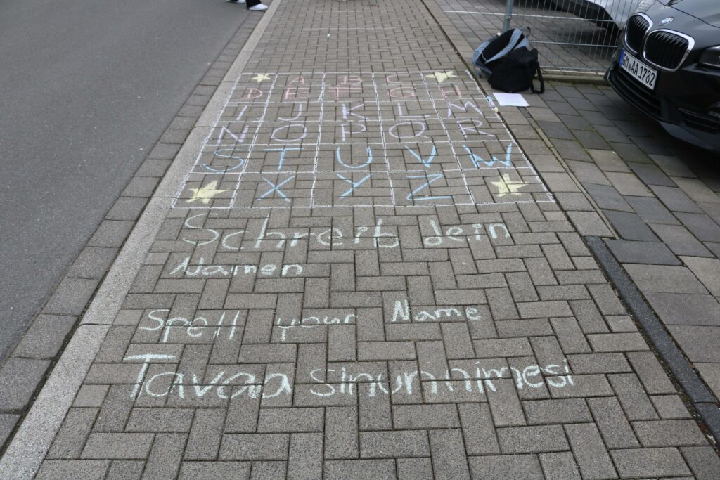 Bunter Straßenmalkreide-Text auf einem Gehweg mit Namen und Spielanleitung.
