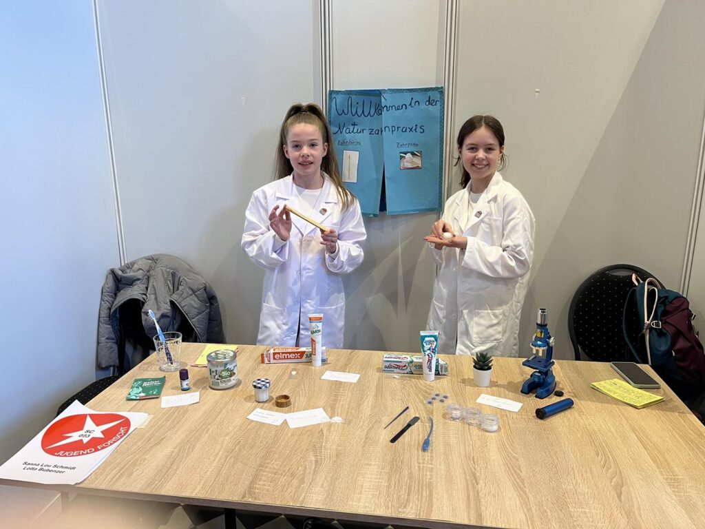 Zwei Schülerinnen in Laborkitteln stehen an einem Tisch mit Experimenten und Materialien.