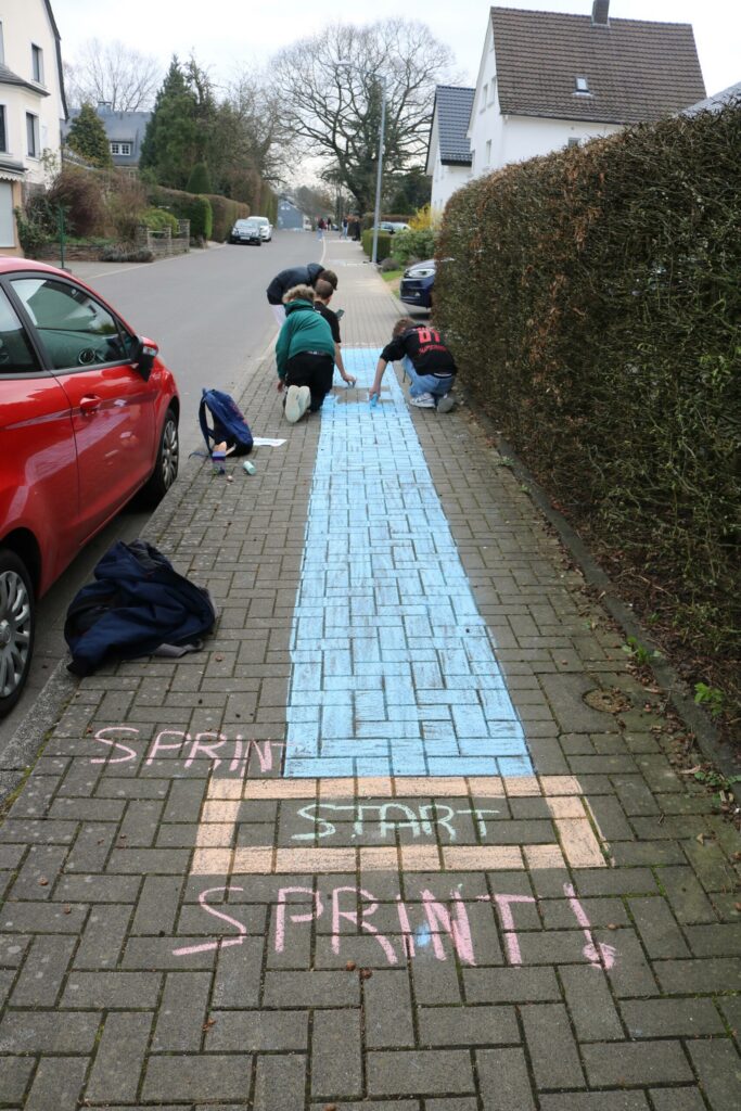 Schüler gestalten einen Sprintrennbahn auf dem Bürgersteig mit Kreide.