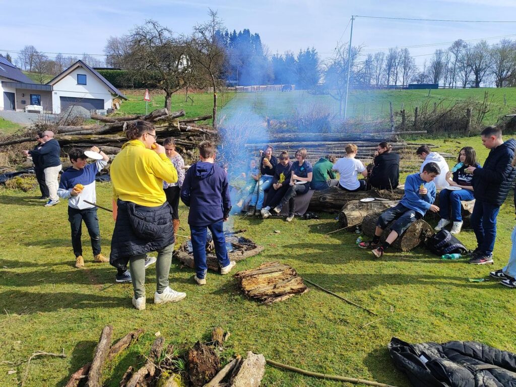 Grupp von Schülern sitzt um ein Feuer im Freien, im Hintergrund Wiese und Bäume.