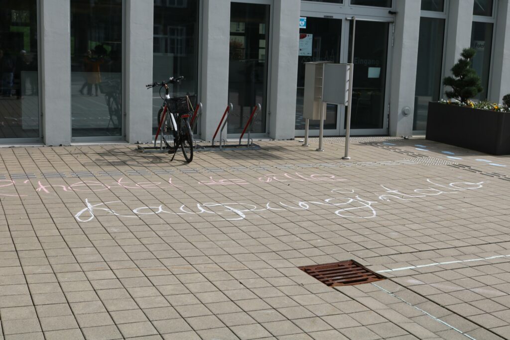 Schriftzug auf dem Boden vor einem Gebäude mit Fahrrad.