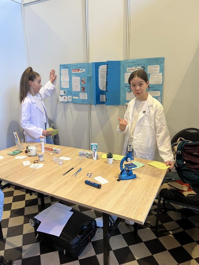 Zwei Schülerinnen in Laborkitteln präsentieren ein Experiment an einem Tisch.
