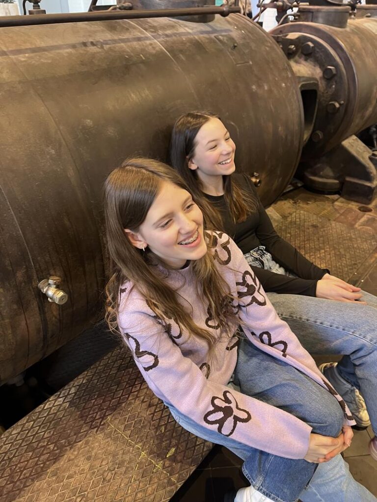 Zwei Mädchen sitzen lächelnd auf einer Holzbank neben einem großen Metallgerät.