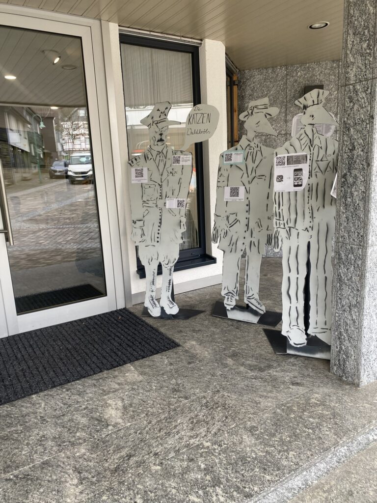Drei lebensgroße Pappfiguren stehen vor einem Eingangsbereich.