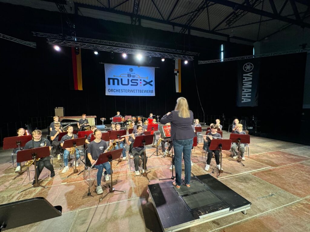 Probeschor mit Musizierenden auf einer Bühne, dirigiert von einer Frau.