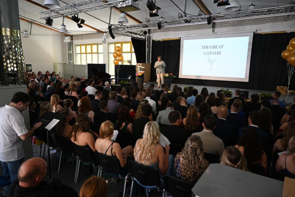 Veranstaltung mit vielen Zuschauern in einem Saal, Bühne und Präsentation im Hintergrund.