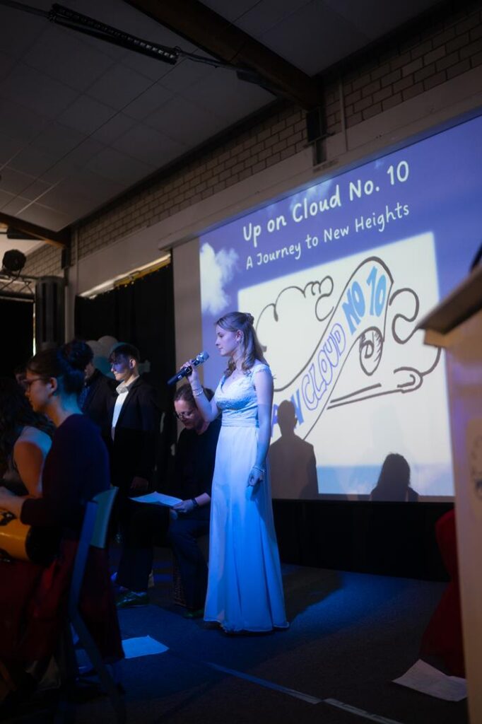 Schülerin in weißem Kleid singt auf einer Bühne mit Projektion im Hintergrund.