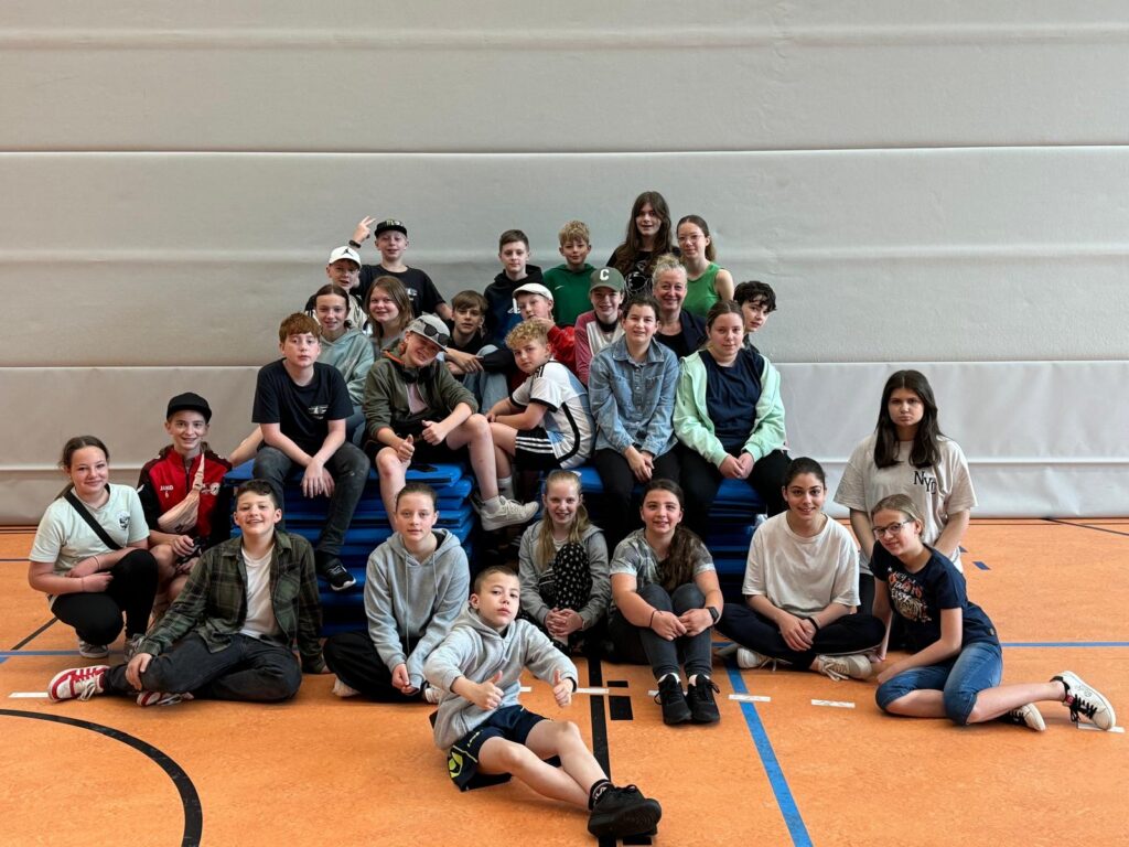 Gruppe von Schülerinnen und Schülern sitzt auf Turnmatten in einer Sporthalle.
