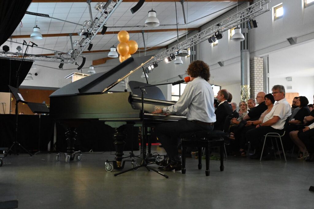Ein Musiker spielt Klavier während einer Veranstaltung; Publikum sitzt aufmerksam im Hintergrund.