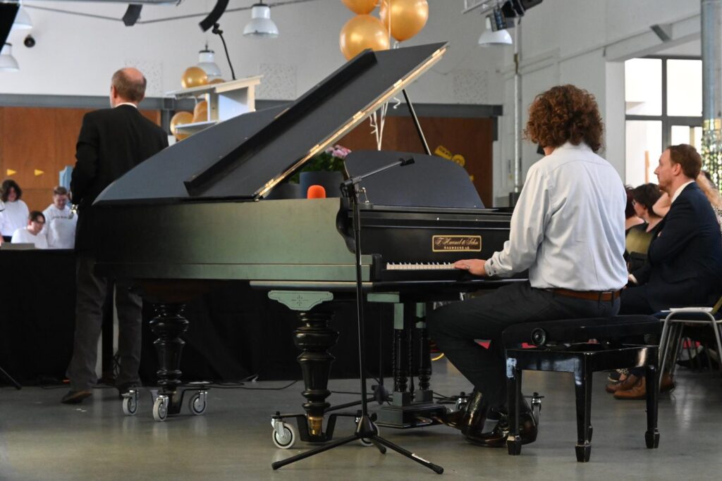 Ein Pianist spielt an einem schwarzen Klavier, während im Hintergrund eine Veranstaltung stattfindet.