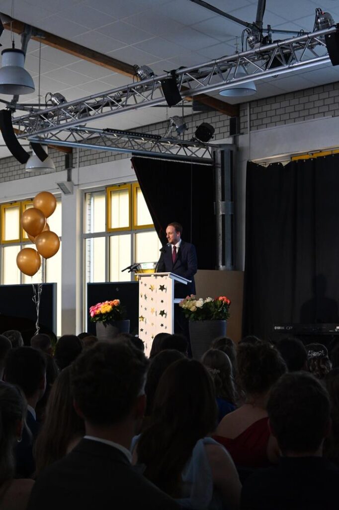 Ein Mann steht am Rednerpult vor einem Publikum in einem festlich dekorierten Raum.