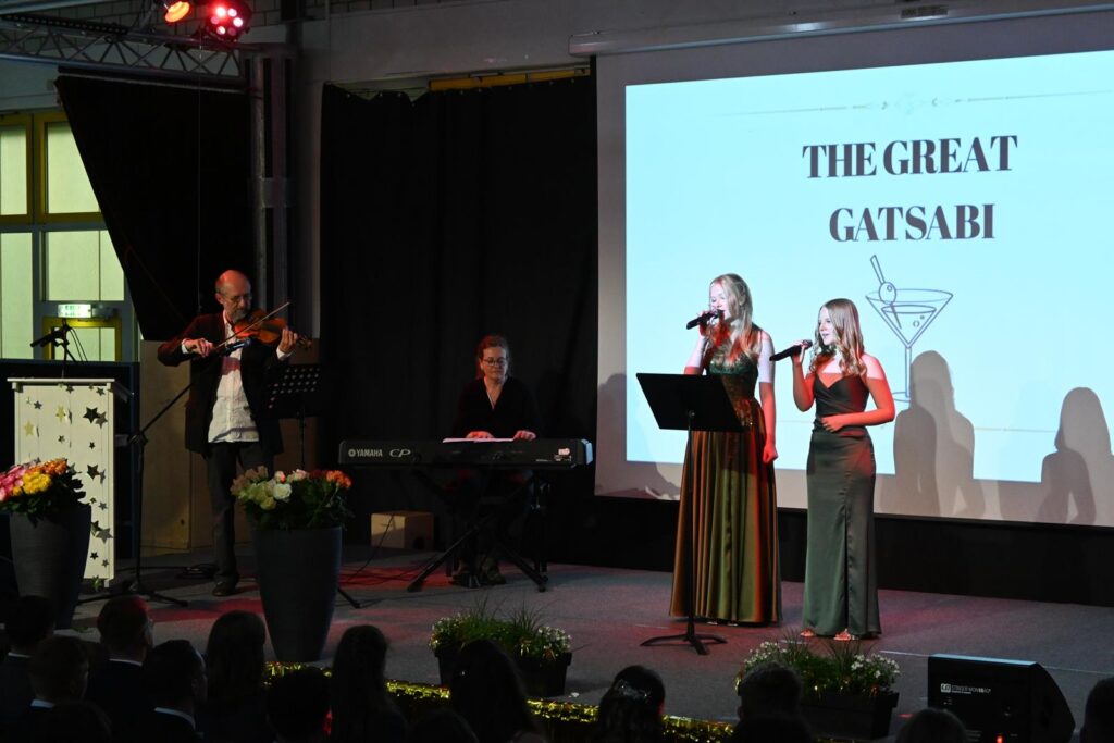 Zwei Sängerinnen auf der Bühne mit einem Musiker und einer Präsentation im Hintergrund.